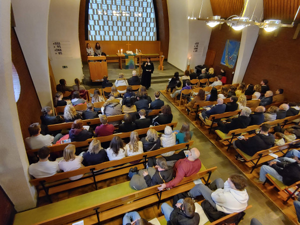 Blick in den Altar- und Kirchraum von oben. Viele Bänke sind voll besetzt. Eine Pfarrerin steht frontal.