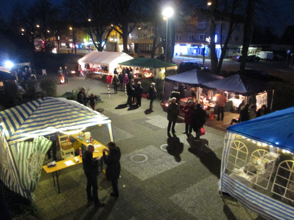 Blick bei Dunkelheit auf den beleuchteten Kirchvorplatz, auf dem Pavillons mit Weihnachtsmarktständen stehen.