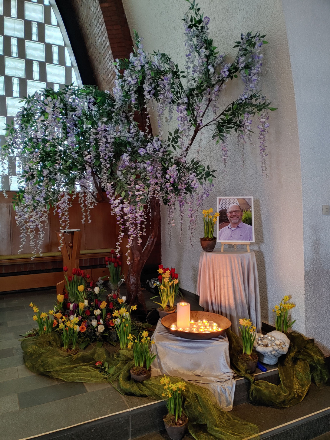 Ein Foto des verstorbenen Pfarrer Martin Gres steht auf einem weiß abgehangenen Tisch. Um ihn herum viele bunte Blüten und davor eine Schale mit vielen brennenden Kerzen.