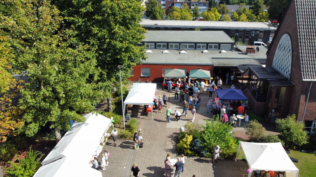 Das Gemeindefest 2024 mit vielen Menschen an den Ständen, an Tischen und Bänken auf dem Kirchvorplatz und dem Parkplatz aus der Luft fotografiert.