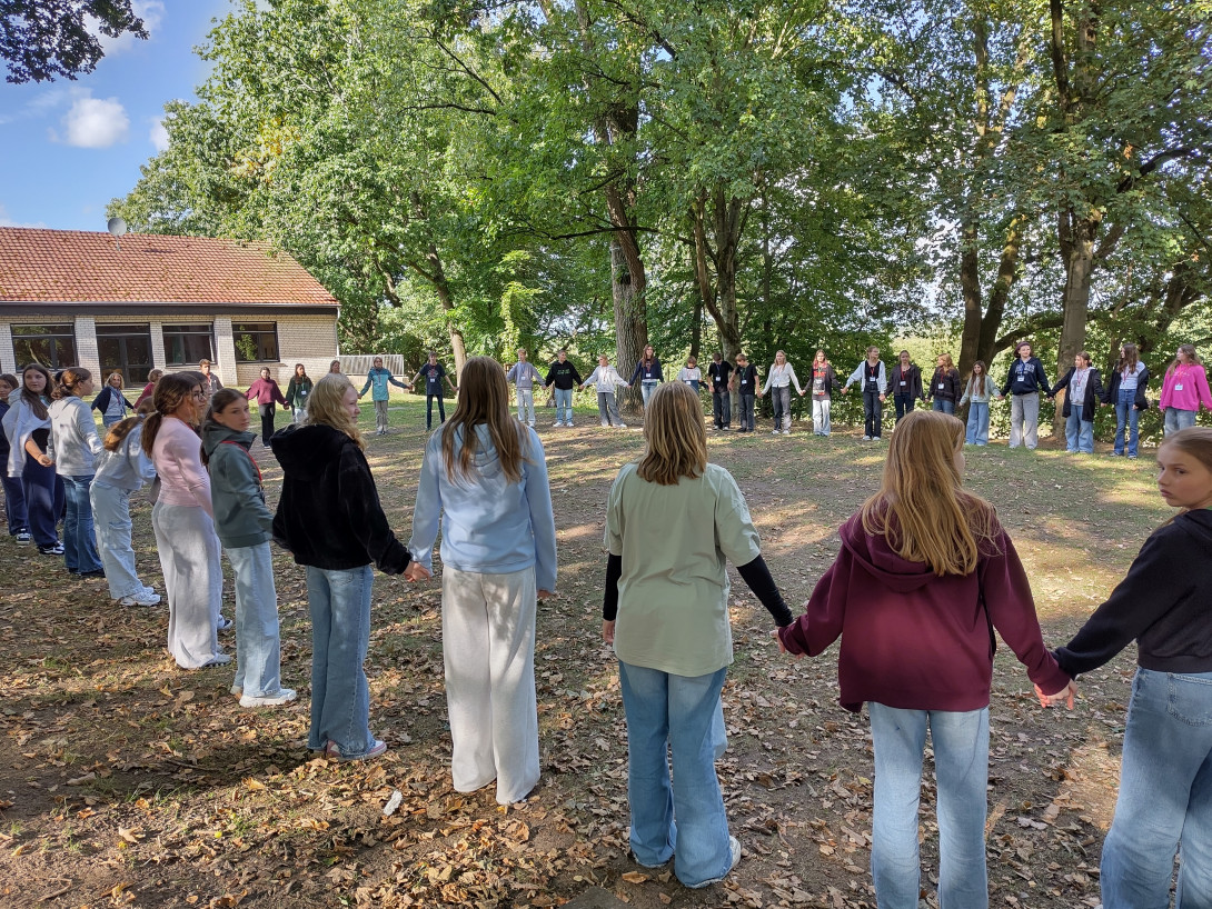 60 Konfis stehen Hand an Hand in spätsommerlichem Grün vor einem Freizeitheim im Kreis.