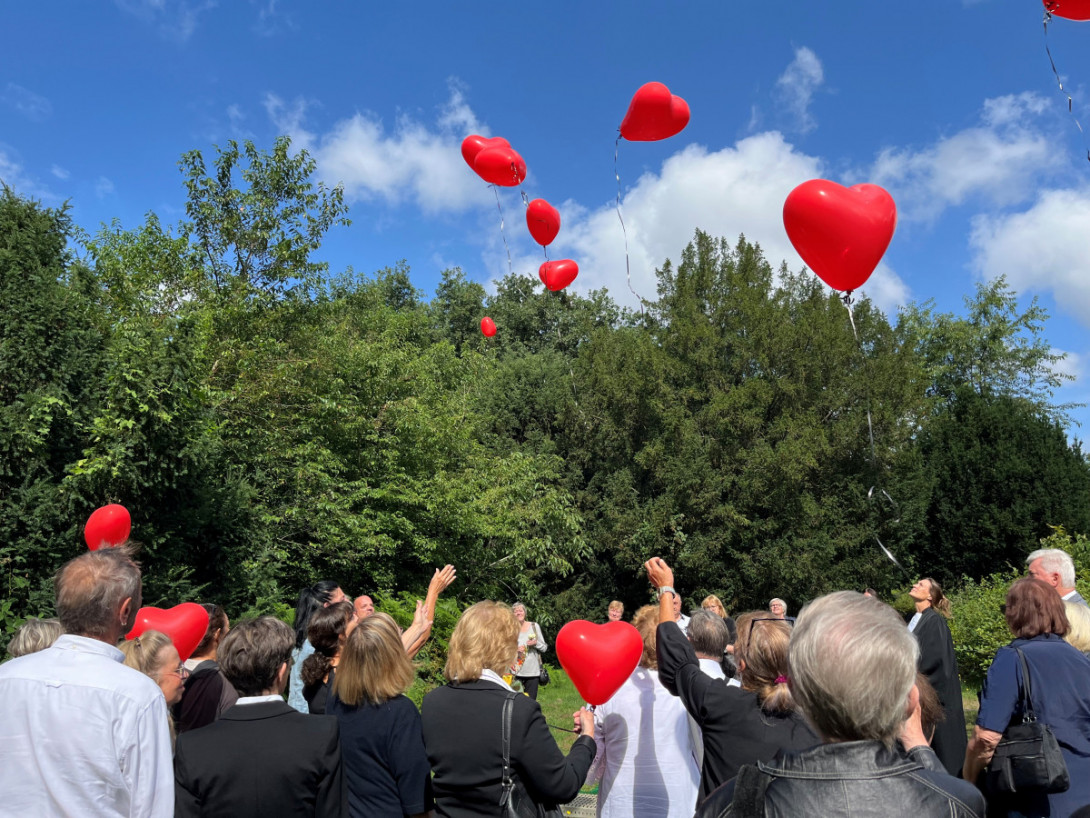 Eine Gruppe von Menschen lässt rote herzförmige Luftballons in den blauen Himmel aufsteigen.